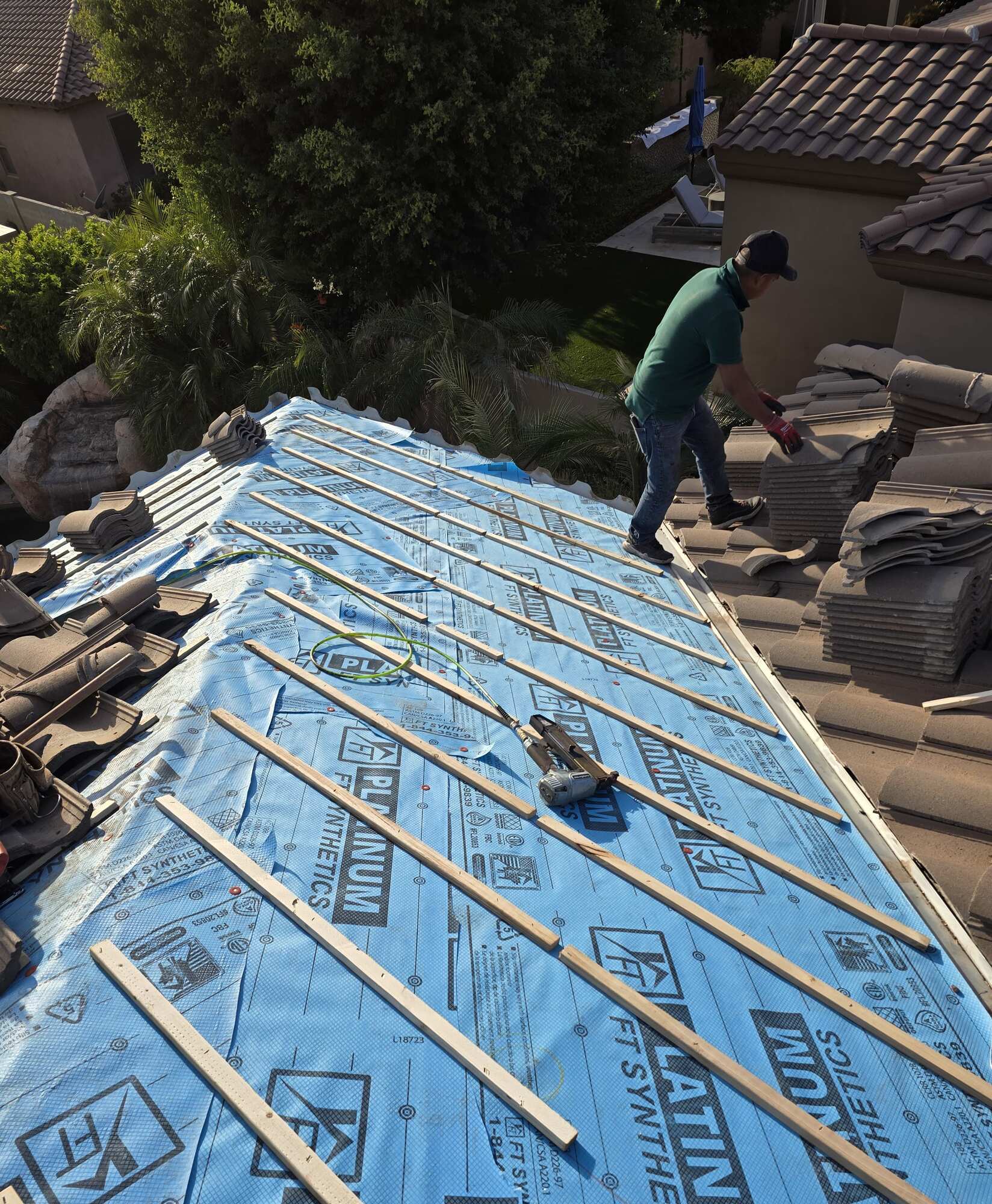 A roofer in a green shirt and cap carries tile across a roof beside a large plane of blue synthetic underlayment battened out and ready for tile installation.
