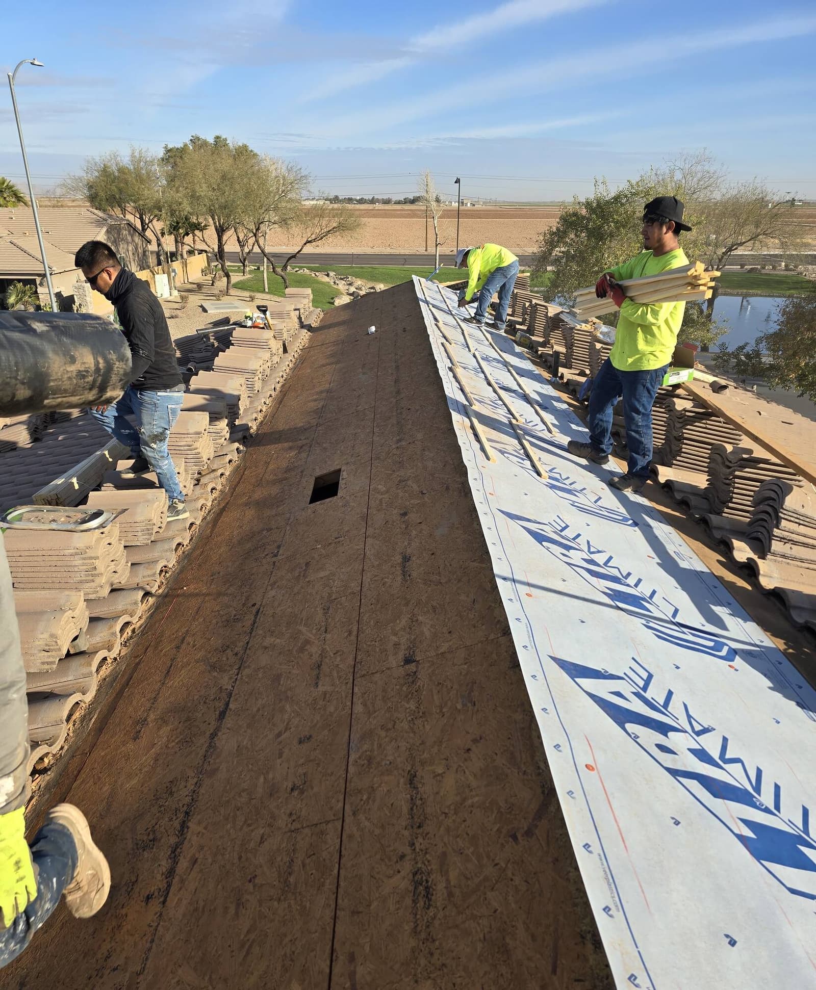 Roofing crew mid-project on a tile re-roof, rolling out underlayment and carrying battens across a stripped deck with salvaged concrete tiles stacked on both slopes.