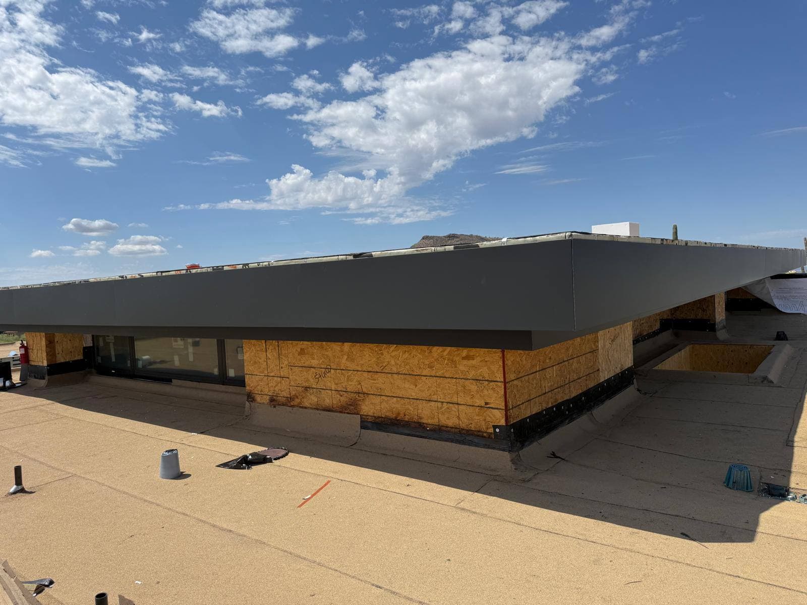 Deep charcoal metal fascia wrapping the roof edge of a modern flat-roof desert home, seen across the tan granulated flat-roof surface.
