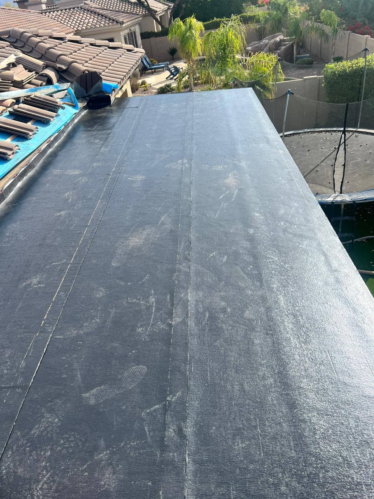 A low-slope roof section covered in new black modified-bitumen membrane, adjoining a tile roof, above a landscaped desert backyard.