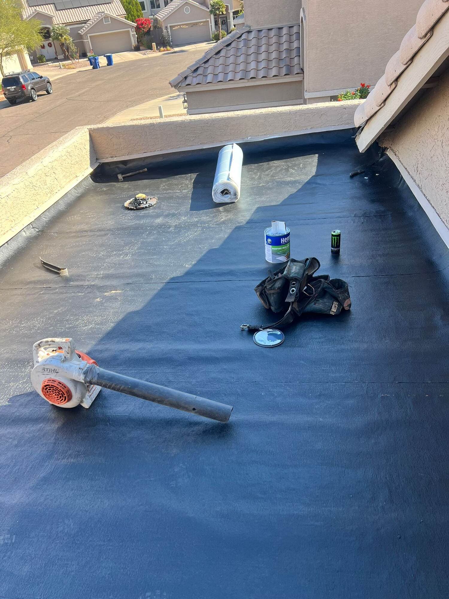 Freshly sealed dark flat roof deck on a Phoenix home, with roofing tools, a blower and a sealant can laid out across the surface and a tile roof section visible beyond the parapet.