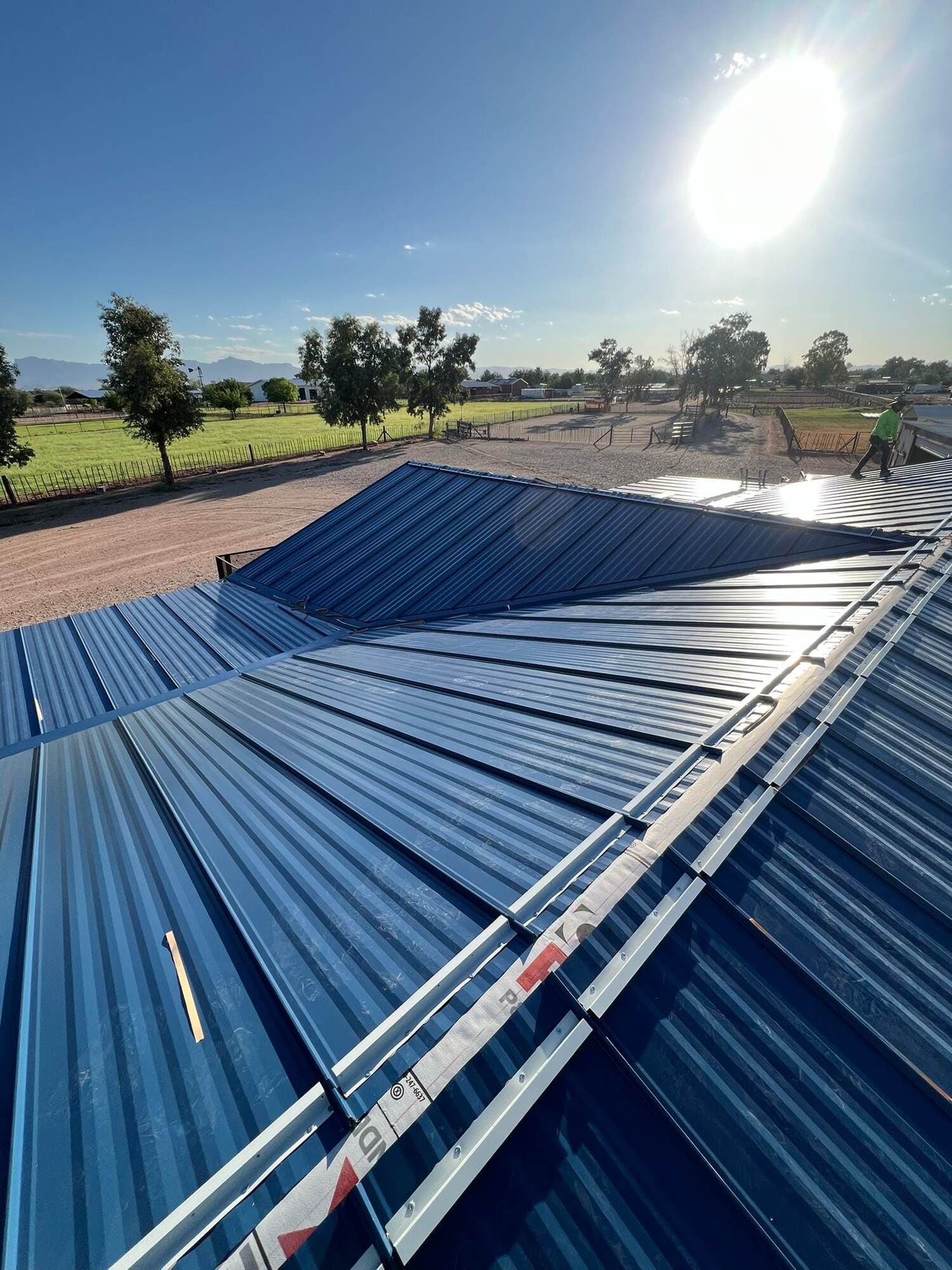 A completed blue metal roof with clean hip and valley lines on a rural Arizona property, with green pasture and distant mountains under a flaring sun.