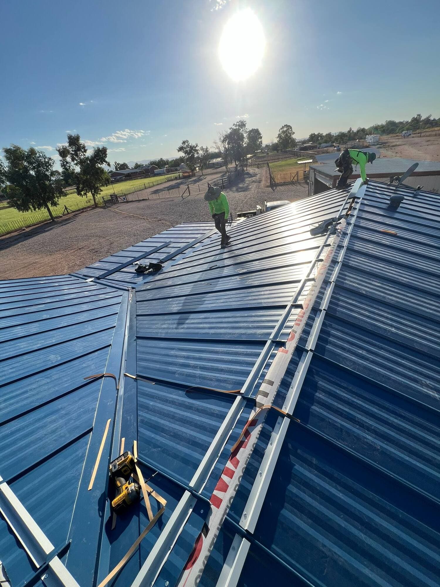 Two roofers in green high-visibility shirts installing a large blue metal roof on a rural barn, with the sun flaring overhead and horse pasture stretching out behind.