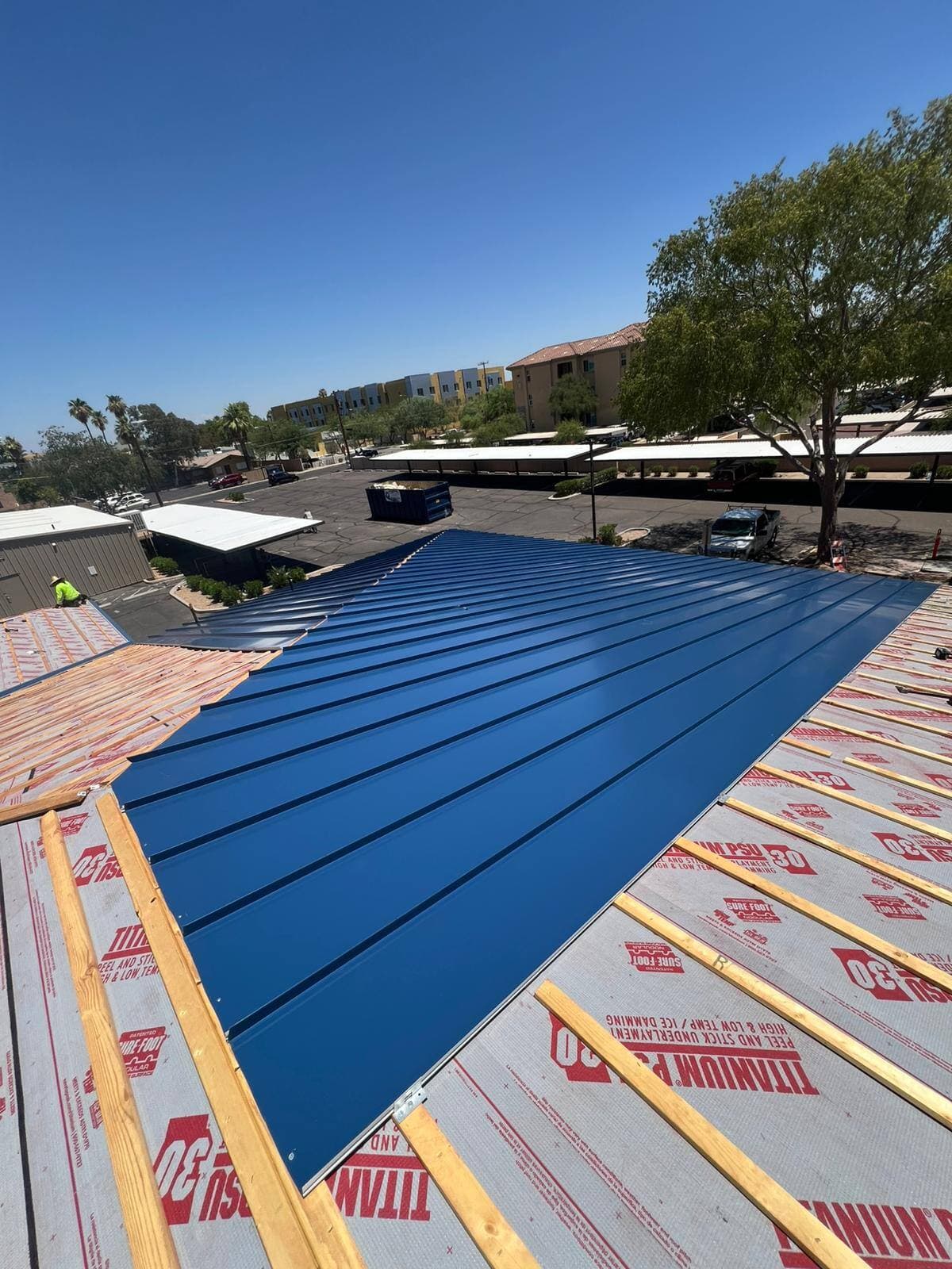 Deep blue standing seam metal panels installed across the center of a commercial roof, flanked by peel-and-stick underlayment and wood battens still awaiting panels.