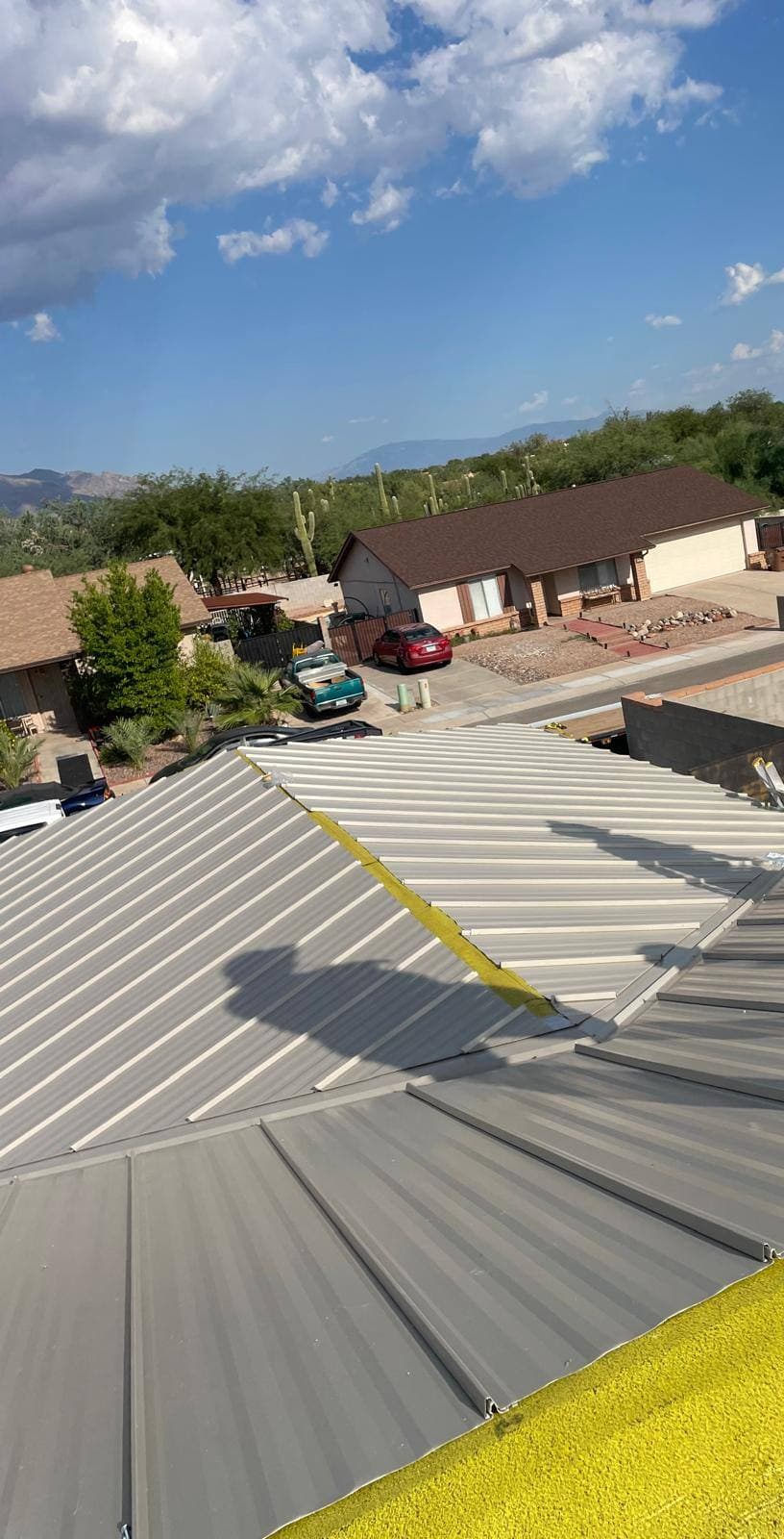 Completed tan standing-seam metal roof with crisp panel lines, looking out over a desert neighborhood to a rugged mountain range on the horizon