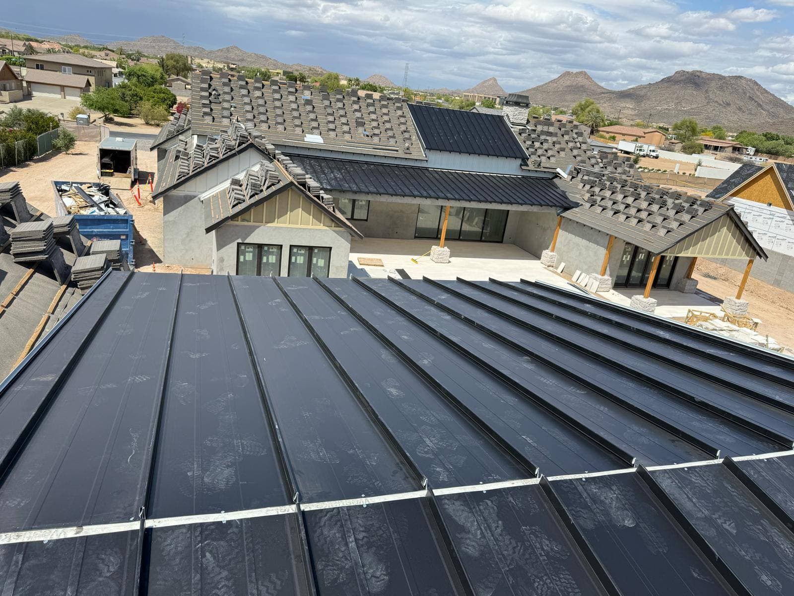 Matte black standing seam metal panels installed on a new desert home while roof tile sits stacked on adjacent slopes, with Arizona mountains behind