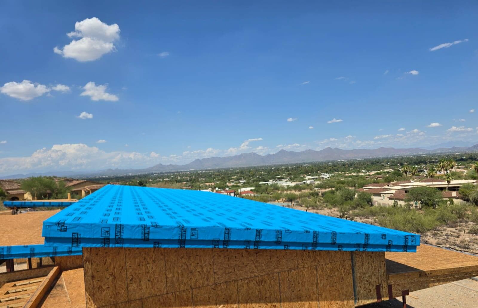 Low-slope new-construction roof dried in with blue self-adhered underlayment, its cantilevered edge fully wrapped, above a wide desert valley view.