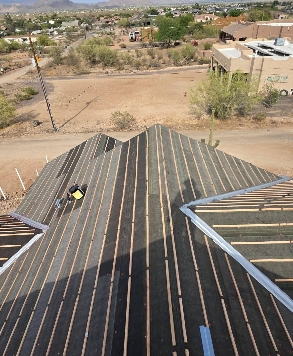 Roof deck covered in dark underlayment with evenly spaced wood battens and new metal valley flashing, seen down the ridge above a desert neighborhood.