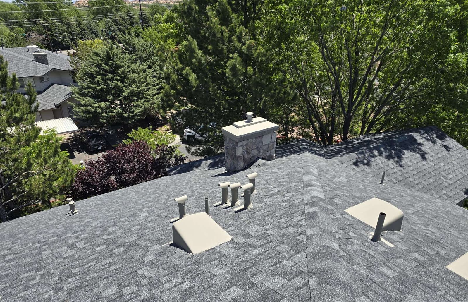 Large finished gray architectural shingle roof with a stone chimney and a bank of color-matched vent stacks, seen from above among mature trees.