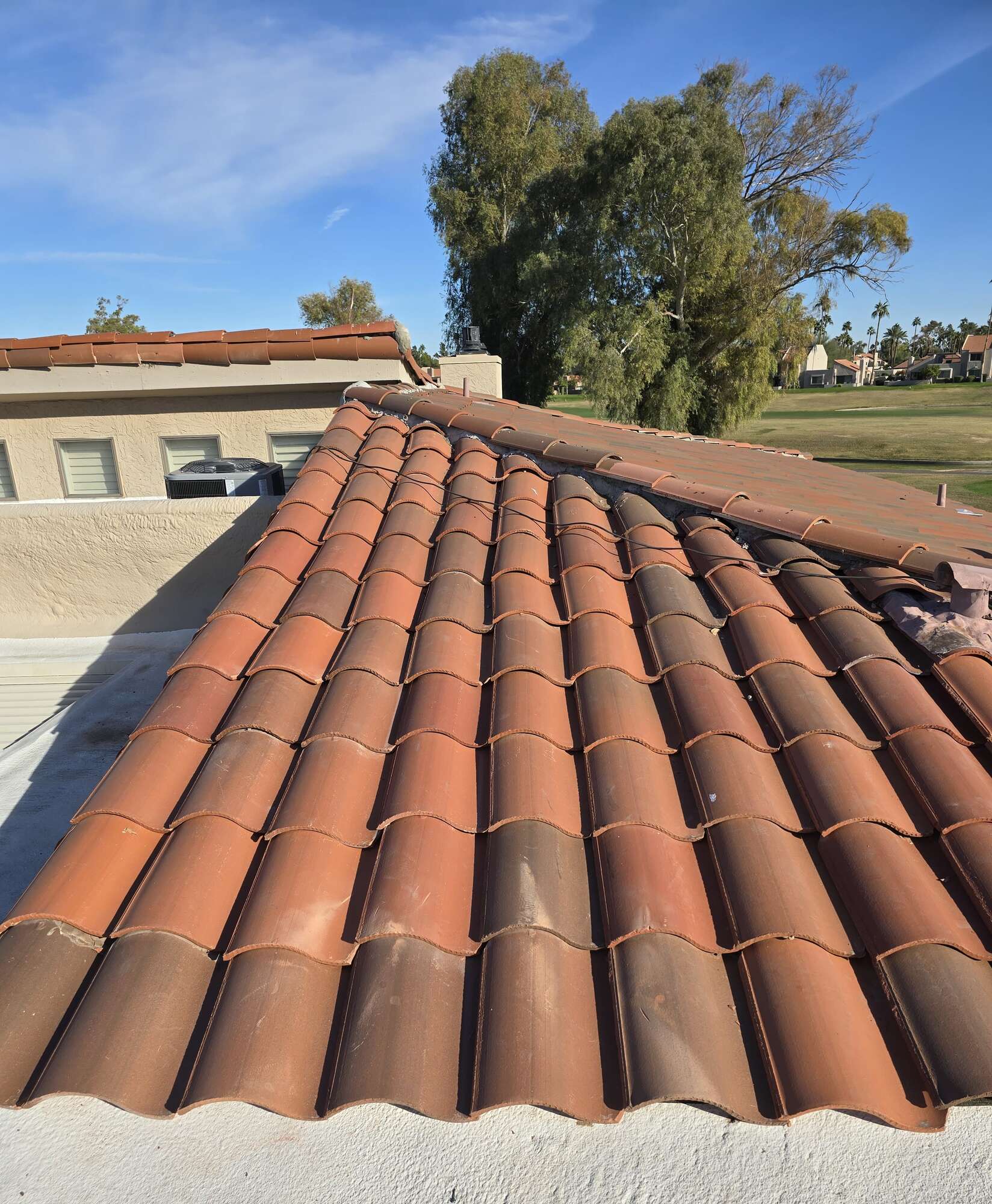 A run of terracotta barrel roof tile leading up to a mortared ridge on an Arizona home, with a golf course fairway and blue sky in the background.