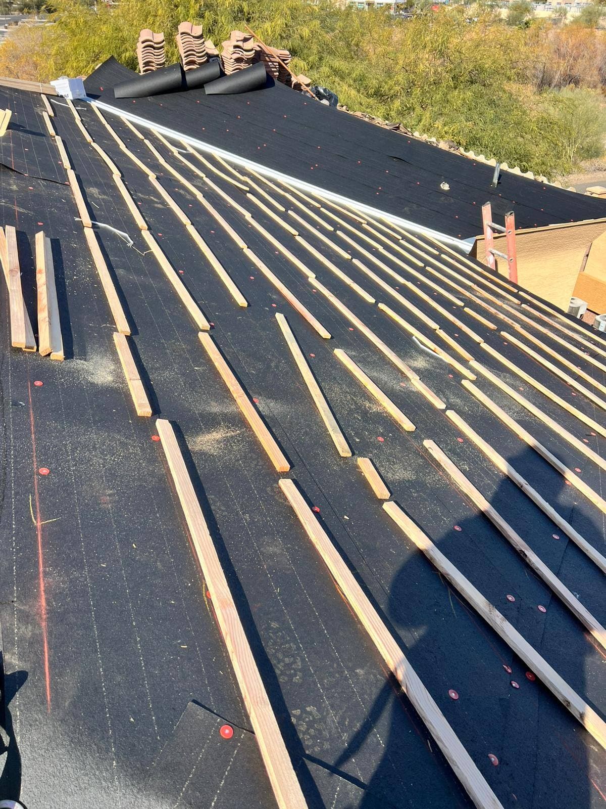 Tile roof in progress with black underlayment, red cap nails and wood battens laid in courses, stacked clay tile at the ridge