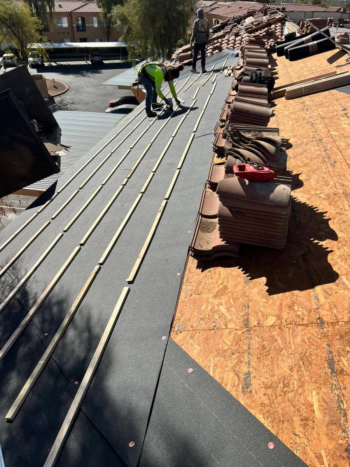 Two roofers working a tile re-roof, one nailing battens over black underlayment while salvaged tiles sit stacked alongside