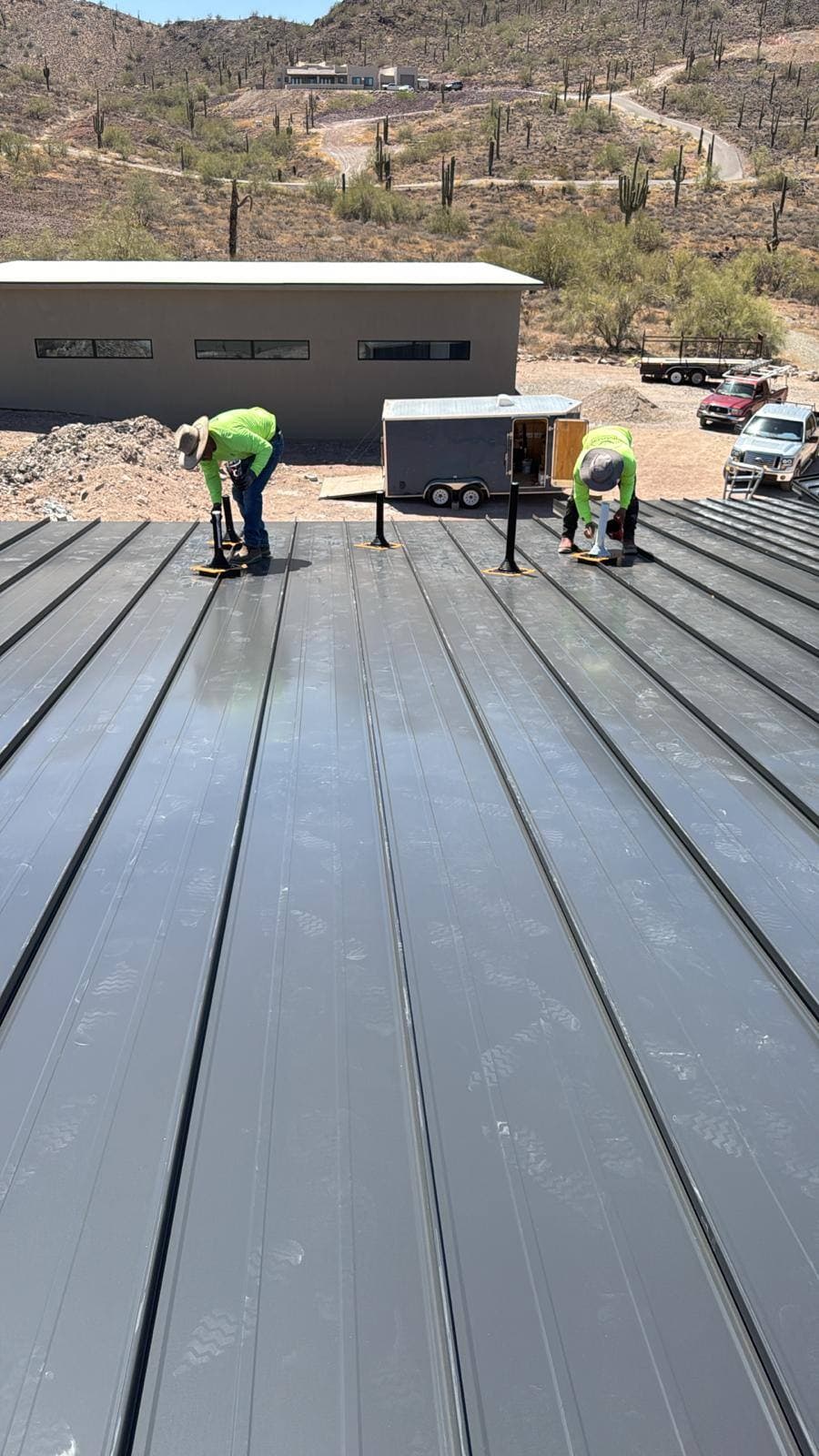 Two roofers in hi-vis shirts and sun hats seaming gray standing seam metal panels on a desert home