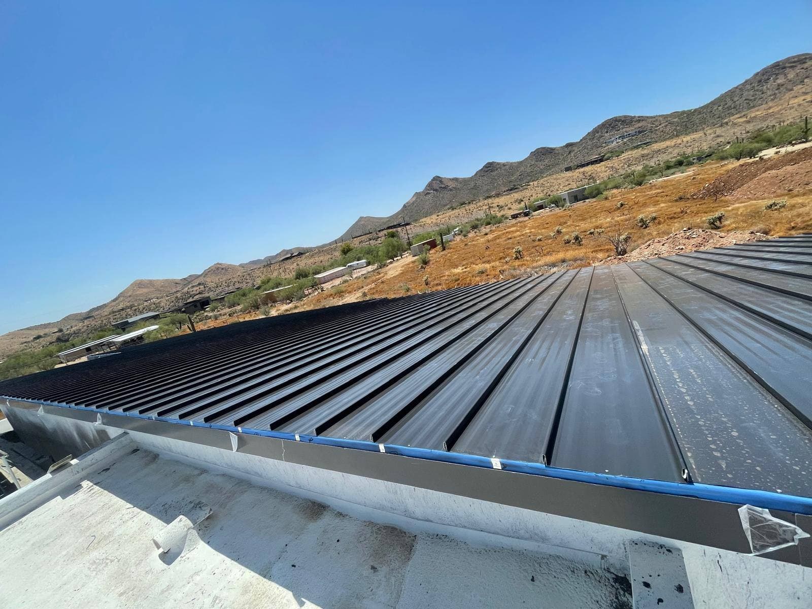 A dark bronze standing seam metal roof with crisp seams, set against a saguaro-covered desert hillside and rocky ridge under clear blue sky.