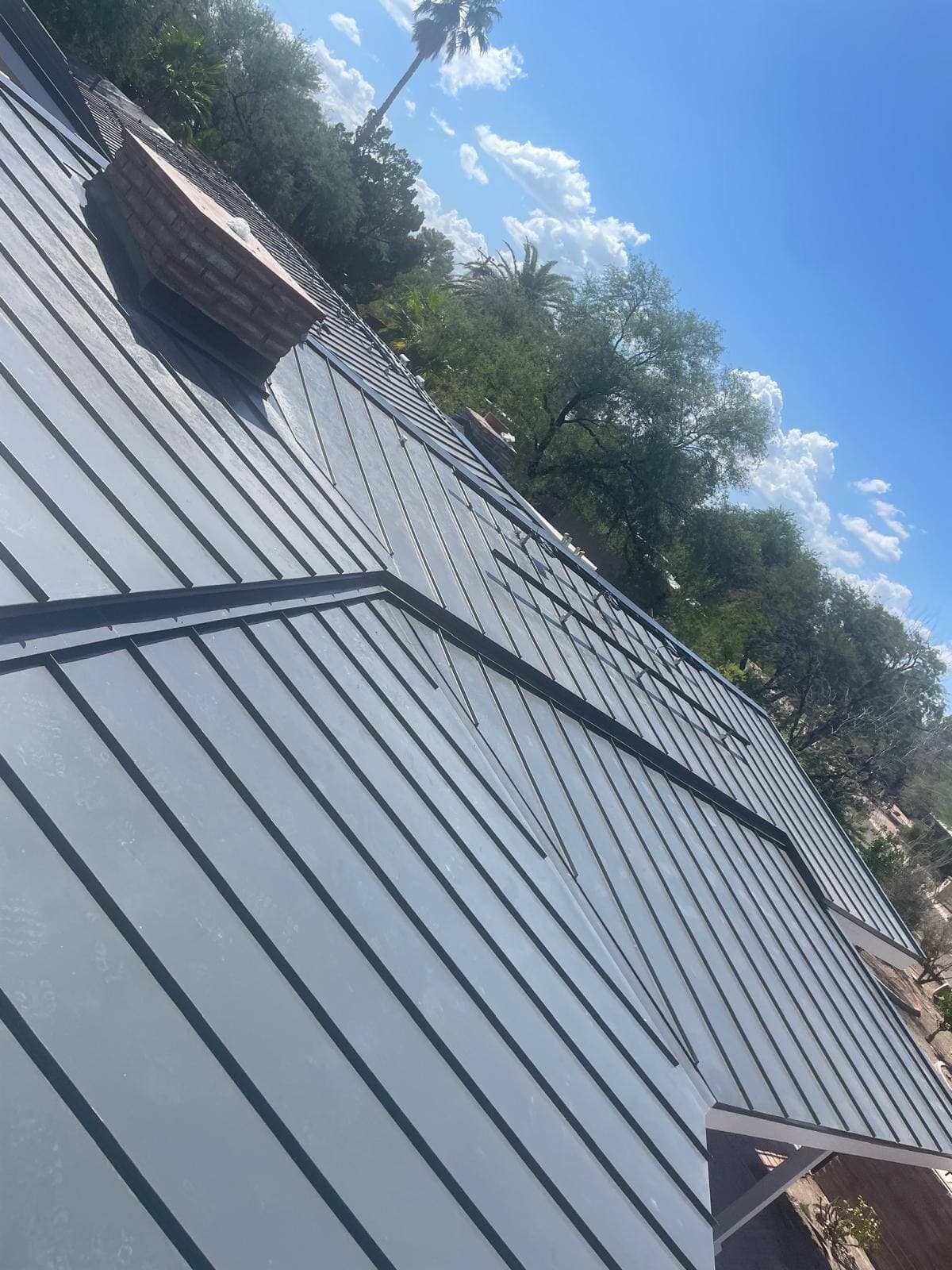 Completed light gray standing-seam metal roof running the length of a large home, with a brick chimney, palms and towering clouds behind