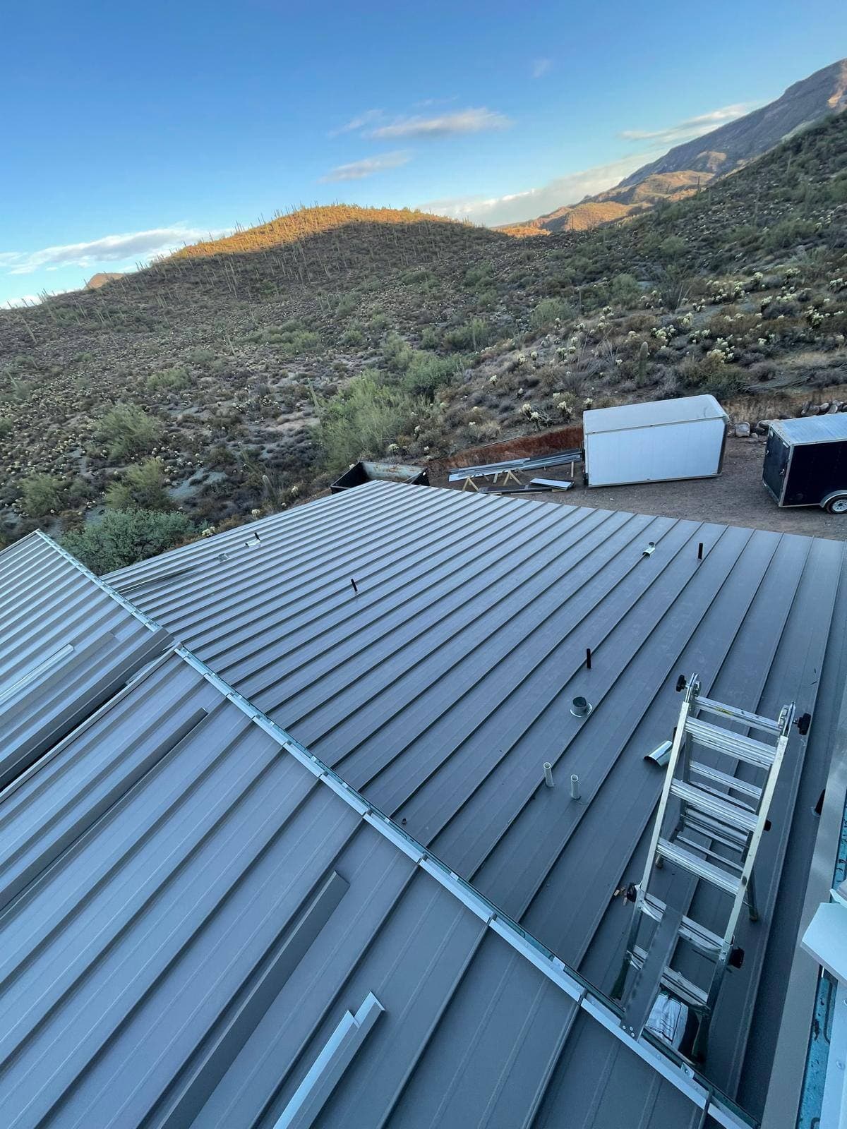 Gray standing-seam metal roof on a desert home with a ladder over the ridge, backed by a saguaro-covered hillside glowing in late-day light