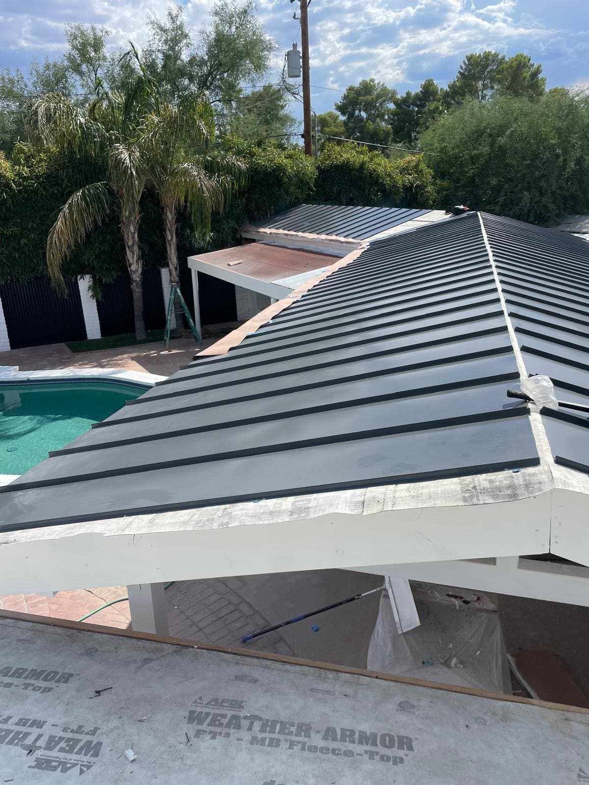 Light gray standing-seam metal roof over a poolside ramada, with a turquoise swimming pool, brick decking and tall palms below a partly cloudy sky