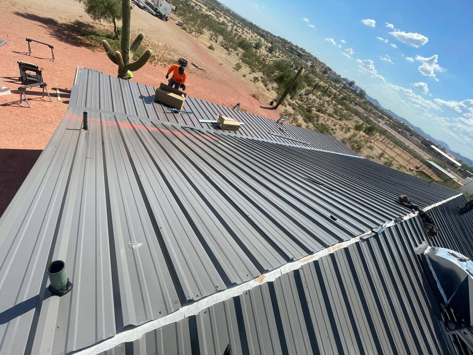 Roofer in orange hi-vis gear working at the ridge of a newly installed gray metal roof, with a saguaro cactus and blue Arizona sky behind