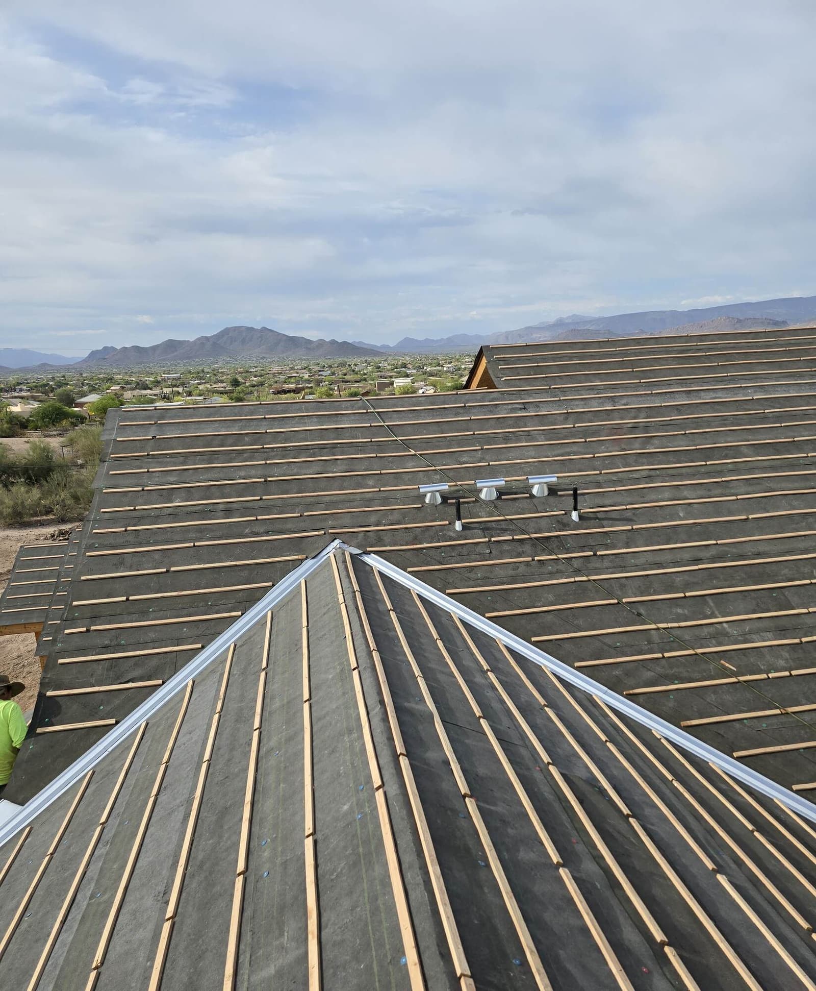 Two converging hip roof planes laid out in dark underlayment with evenly spaced wood battens and bright new metal hip caps, above a desert valley and mountains.