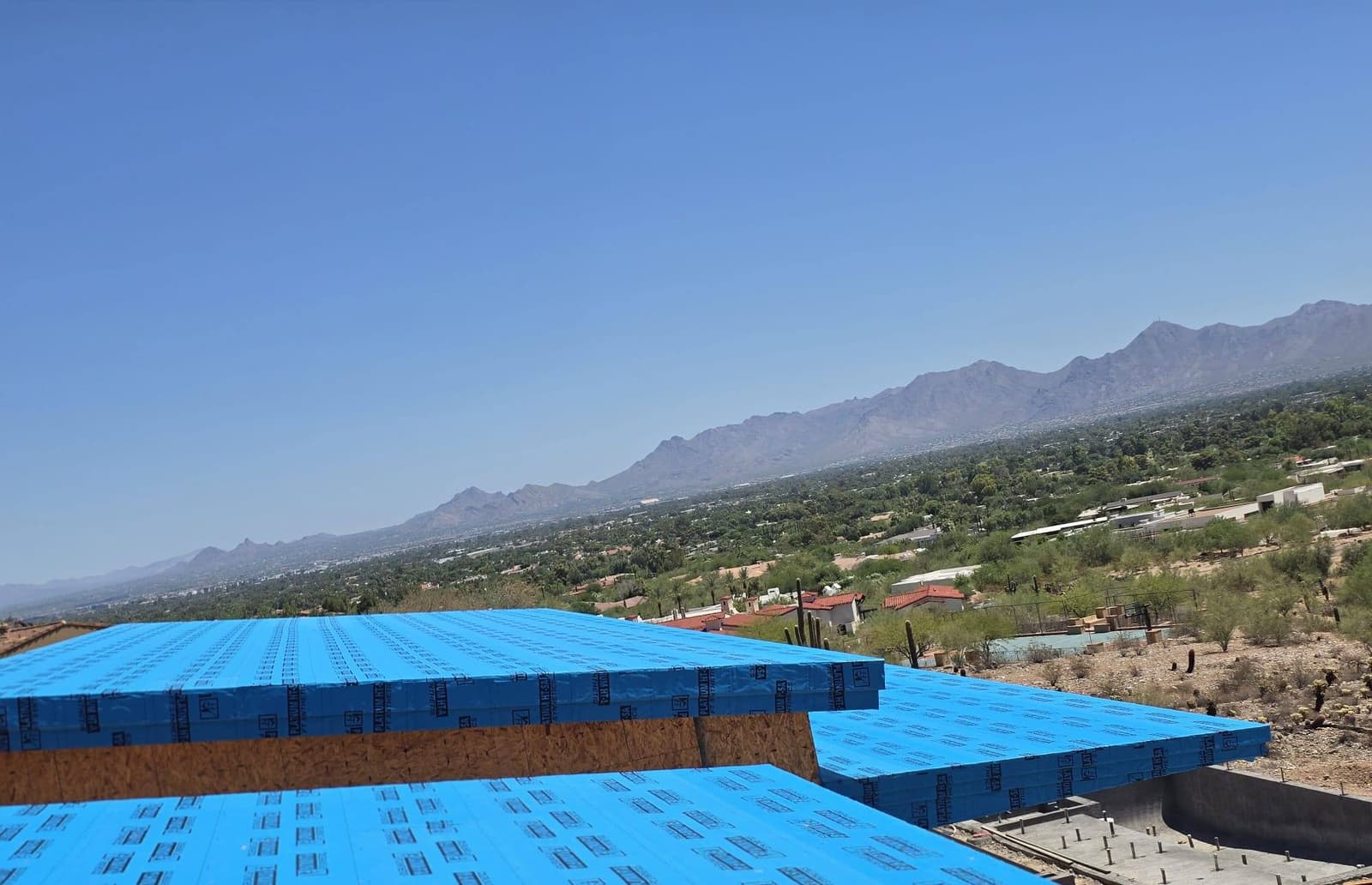 Two stepped flat roof planes dried in with blue synthetic underlayment on a modern hillside home, with a mountain range and valley stretching behind.
